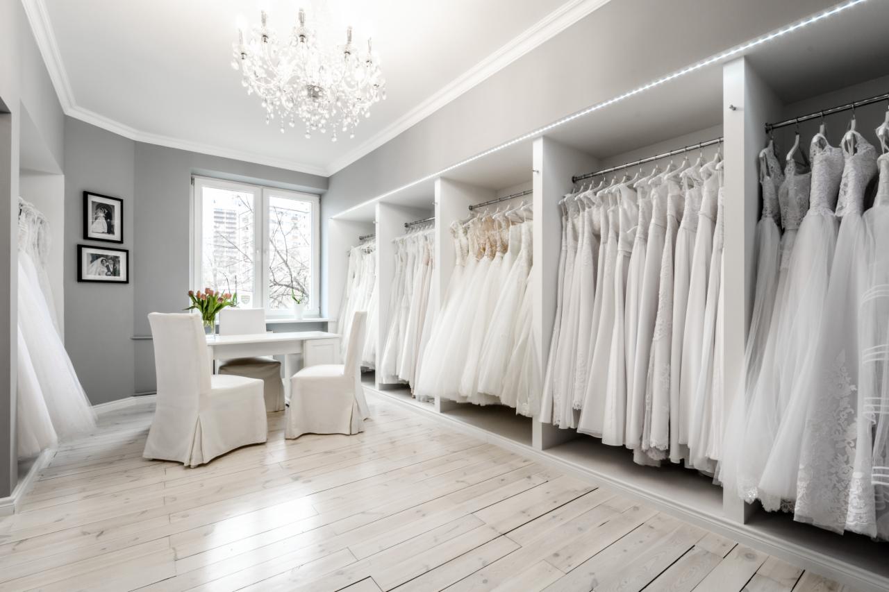 Salon z kolekcją białych sukien ślubnych, podświetlane półki, kryształowy żyrandol, białe meble i bukiet tulipanów na stole.