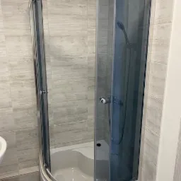 Nowoczesna, narożna kabina prysznicowa z przesuwnymi drzwiami, wykończona chromowanymi detalami, na tle płytek imitujących drewno w odcieniach szarości.