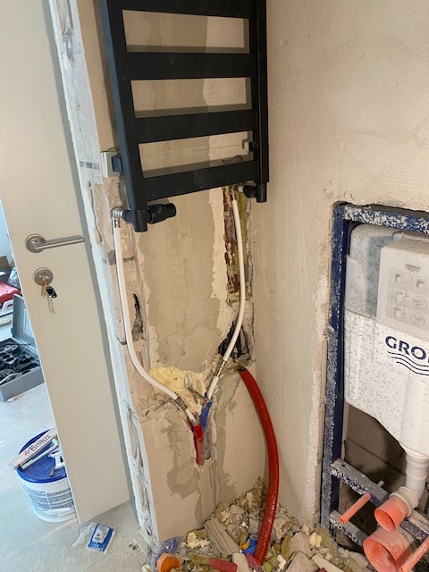 Montaż czarnego grzejnika drabinkowego w łazience podczas prac wykończeniowych, widoczne wystające rury instalacyjne, fragment ściany z uszkodzonym tynkiem i gruzem na podłodze, obok drzwi...