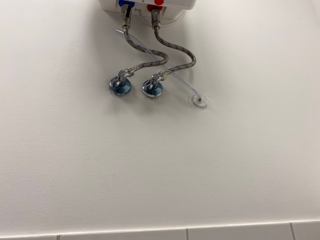 Biała ściana z podłączonym bojlerem elektrycznym, widoczne chromowane zawory, wężyki w oplocie oraz otwór w ścianie z uszczelnieniem silikonowym.