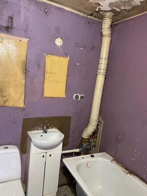 Łazienka w trakcie remontu: fioletowe ściany z żółtymi łatami, biała umywalka z szafką, fragment toalety i wanny, widoczne rury i instalacje, ślady wilgoci na suficie.