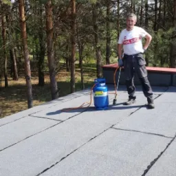 Pracownik w koszulce z napisem 'POLSKA' na dachu pokrytym papą, obok butla gazowa i palnik. W tle drzewa.