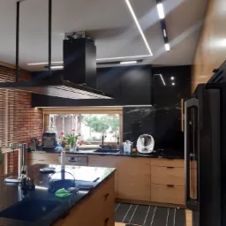 Nowoczesna kuchnia z wyspą, oświetleniem LED podszafkowym i sufitowym, oraz czarnym okapem wiszącym nad płytą, widoczne gniazdka elektryczne w blacie wyspy.