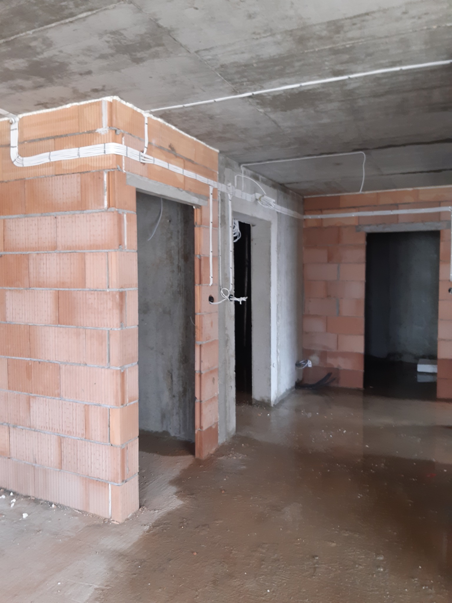 Wnętrze w trakcie budowy z widocznymi przewodami elektrycznymi prowadzonymi po ścianach z czerwonej cegły i pod sufitem, surowy betonowy strop, mokra podłoga.