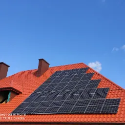 Czerwony dach z dachówki ceramicznej, na którym zamontowano panele słoneczne; widoczne kominy i okno dachowe w trakcie budowy, na tle błękitnego nieba.