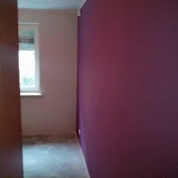 Pokój po pomalowaniu jednej ściany na kolor śliwkowy, druga ściana w odcieniu beżu, widoczne ślady farby na podłodze.