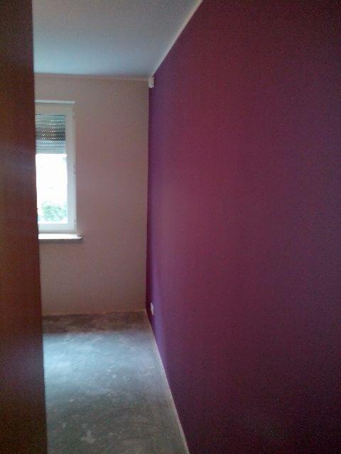 Pokój po pomalowaniu jednej ściany na kolor śliwkowy, druga ściana w odcieniu beżu, widoczne ślady farby na podłodze.