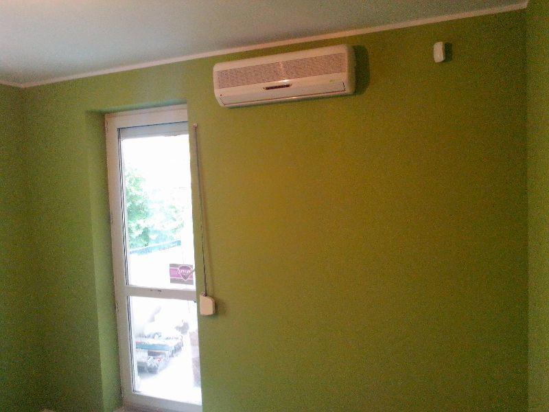 Pokój po odświeżeniu ścian farbą w odcieniu oliwkowej zieleni, widoczny klimatyzator na ścianie oraz okno z białymi ramami.