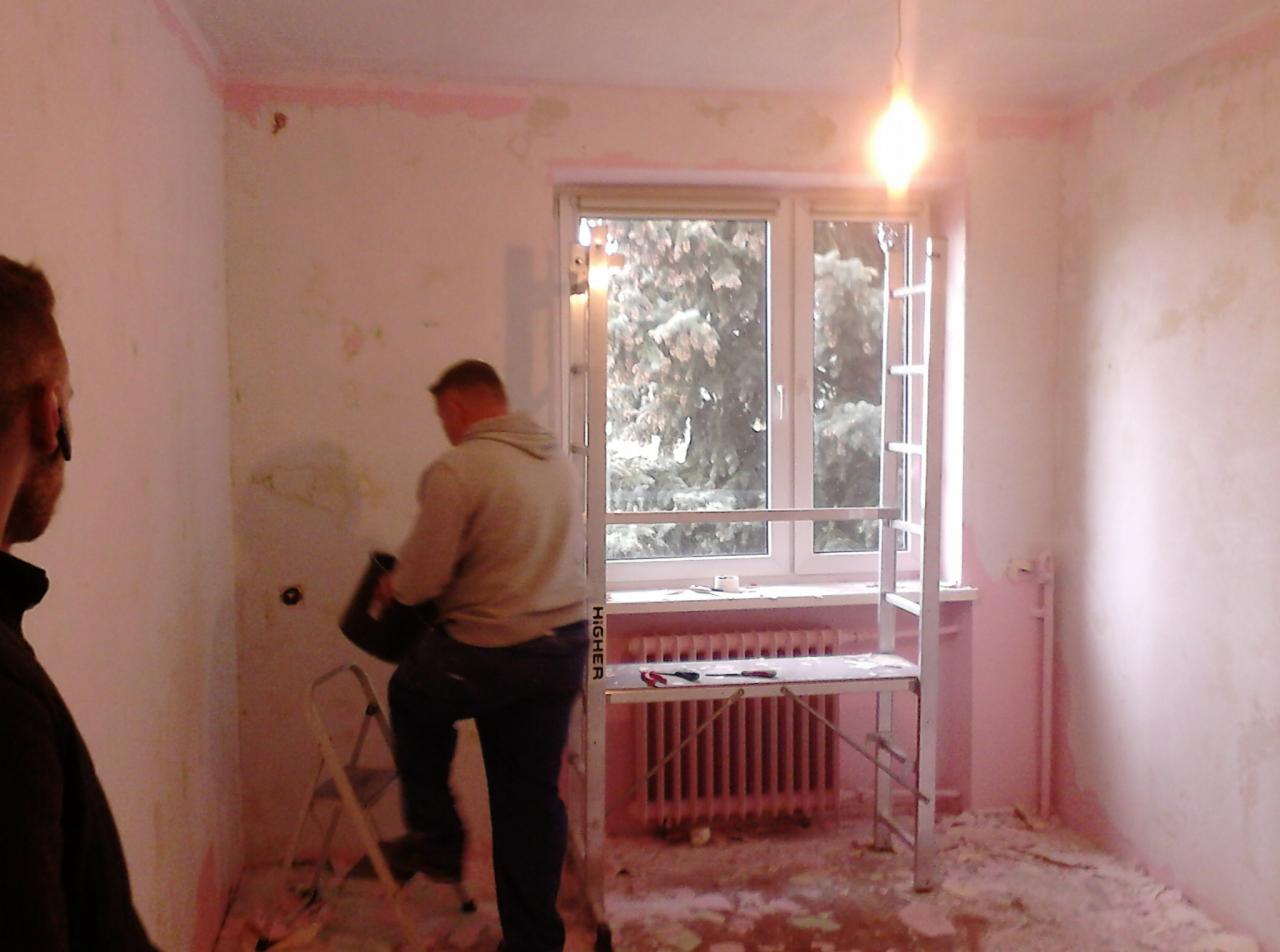 Pokój w trakcie przygotowania do malowania: ściany z resztkami starej farby, drabina, rusztowanie, narzędzia malarskie, widoczne pozostałości po skrobaniu ścian na podłodze.