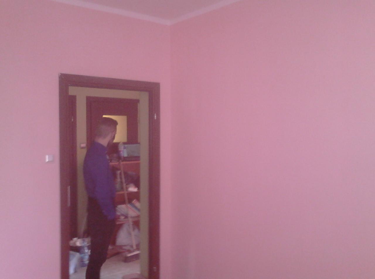Różowa ściana w pokoju po świeżym pomalowaniu, widoczny fragment osoby stojącej w drzwiach, wnętrze z półkami w tle.