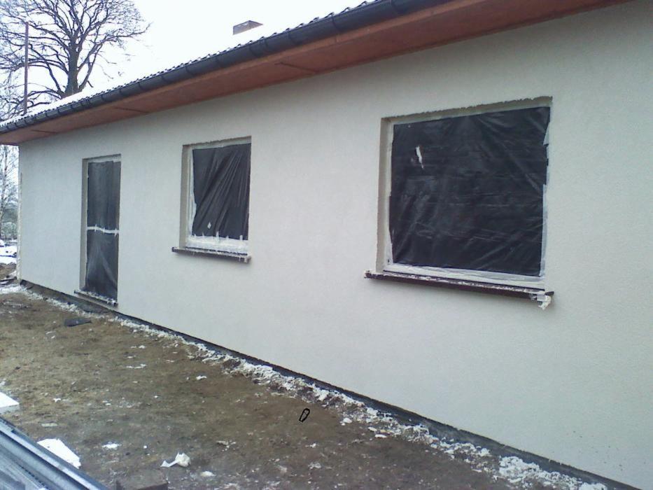 Świeżo otynkowana elewacja budynku jednorodzinnego z zabezpieczonymi folią oknami i drzwiami, widoczne ślady prac wykończeniowych na gruncie.