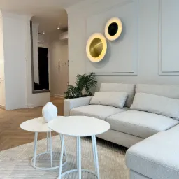 Jasne wnętrze salonu z narożną sofą, dwoma białymi okrągłymi stolikami kawowymi, dekoracyjnymi lampami ściennymi i parkietem ułożonym we wzór jodełki.