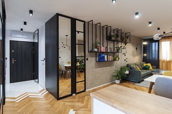 Nowoczesne wnętrze mieszkania z parkietem w jodełkę, czarnymi elementami zabudowy, regałem z książkami i roślinami oraz zieloną sofą z kolorowymi poduszkami.