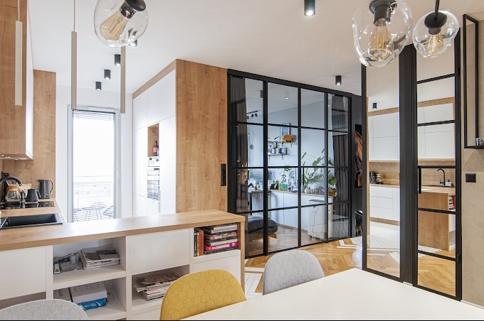 Nowoczesna kuchnia z drewnianymi akcentami i przeszkleniami w czarnej ramie, widok na drugą kuchnię, designerskie lampy wiszące.
