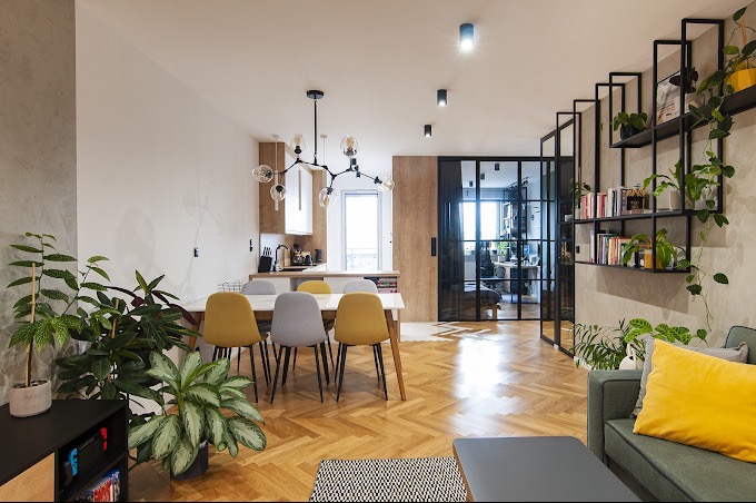 Jasne wnętrze mieszkania w Wołowie z parkietem w jodełkę, nowoczesnym oświetleniem i metalową ścianką działową z półkami na książki i rośliny.