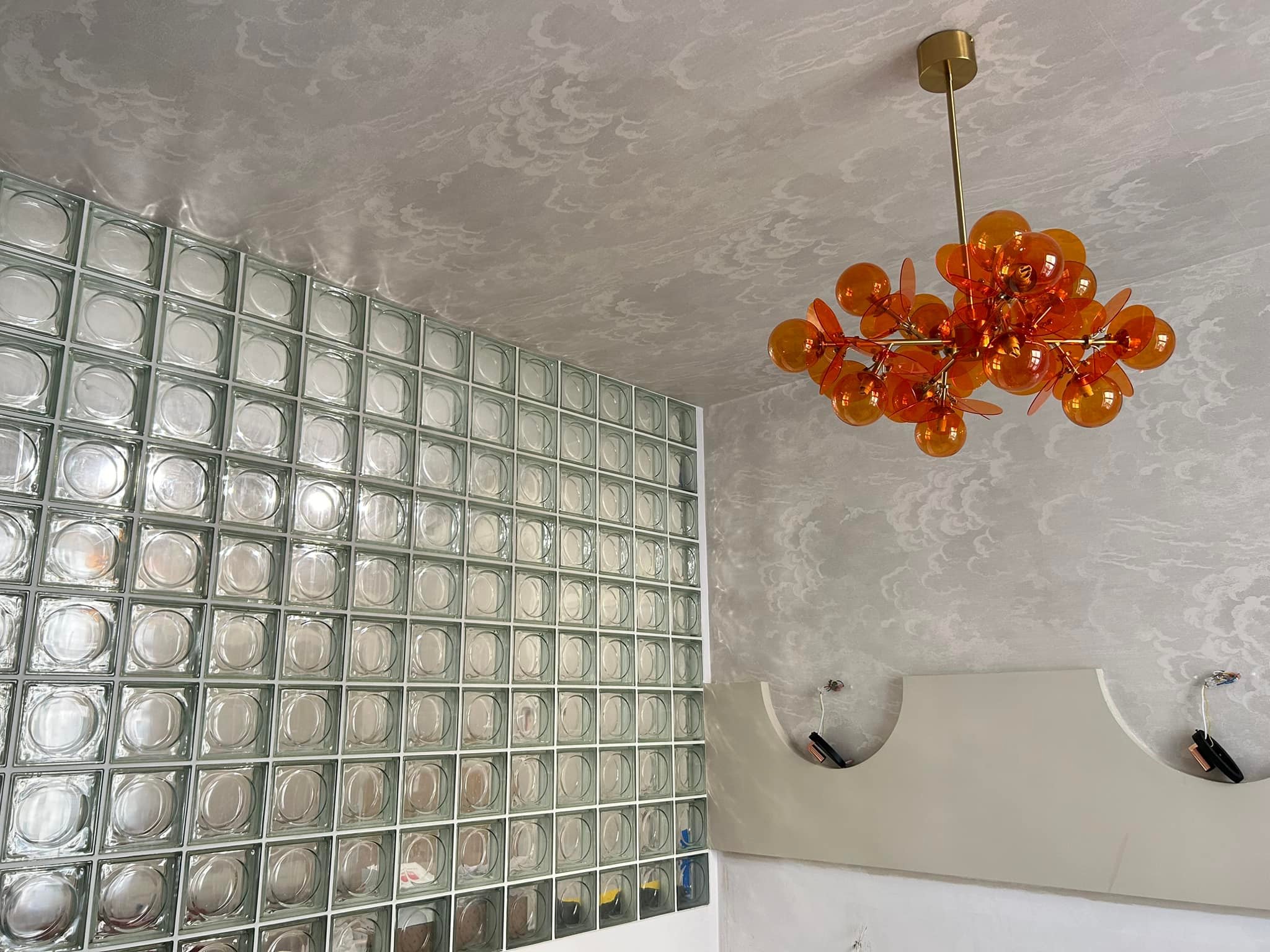 Ściana z luksferów w łazience, oświetlona pomarańczową, designerską lampą wiszącą na tle wzorzystego sufitu.