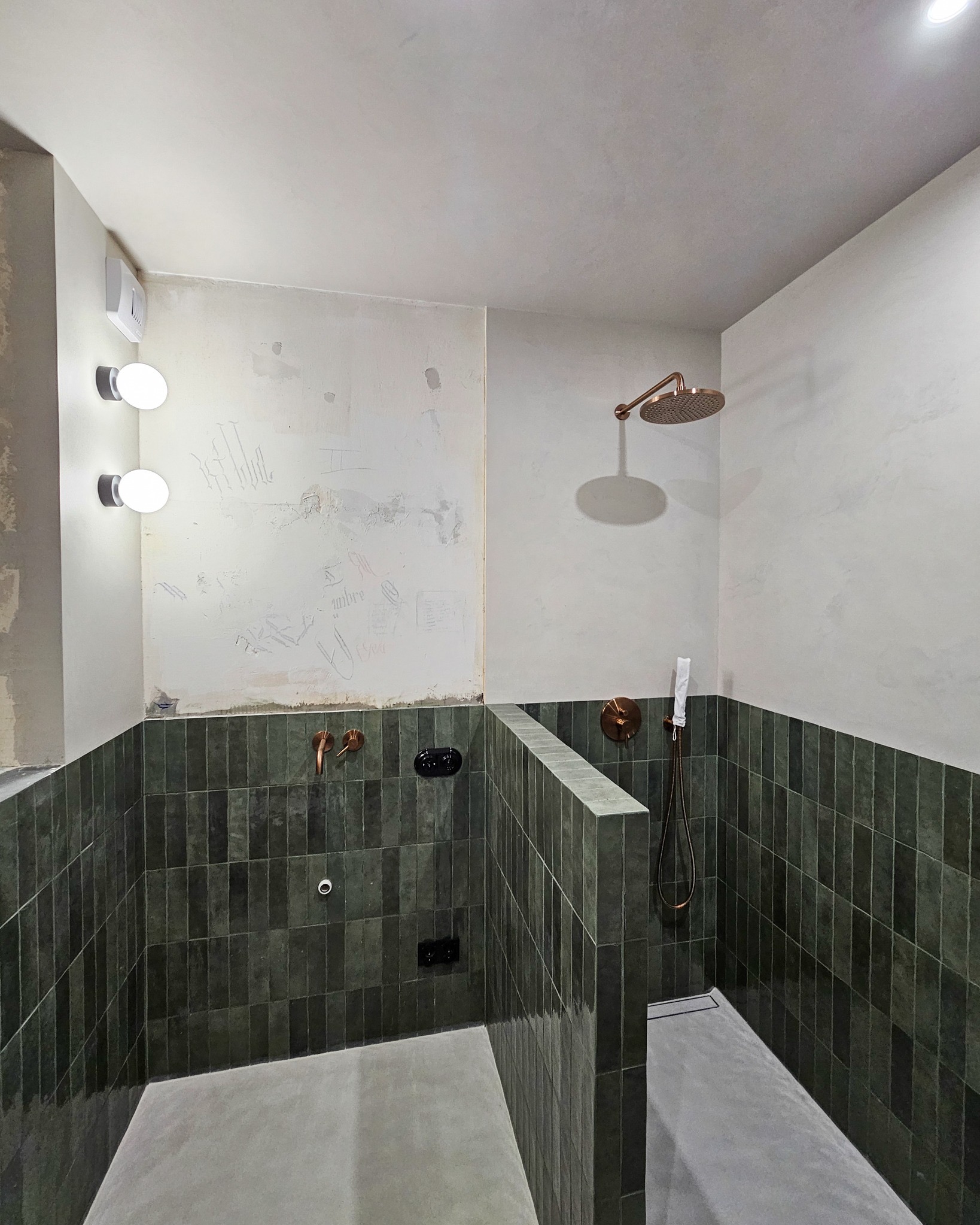 Wnętrze łazienki w trakcie remontu, z zielonymi, pionowo ułożonymi płytkami, miedzianą armaturą i surową ścianą z widocznymi śladami po pracach wykończeniowych.