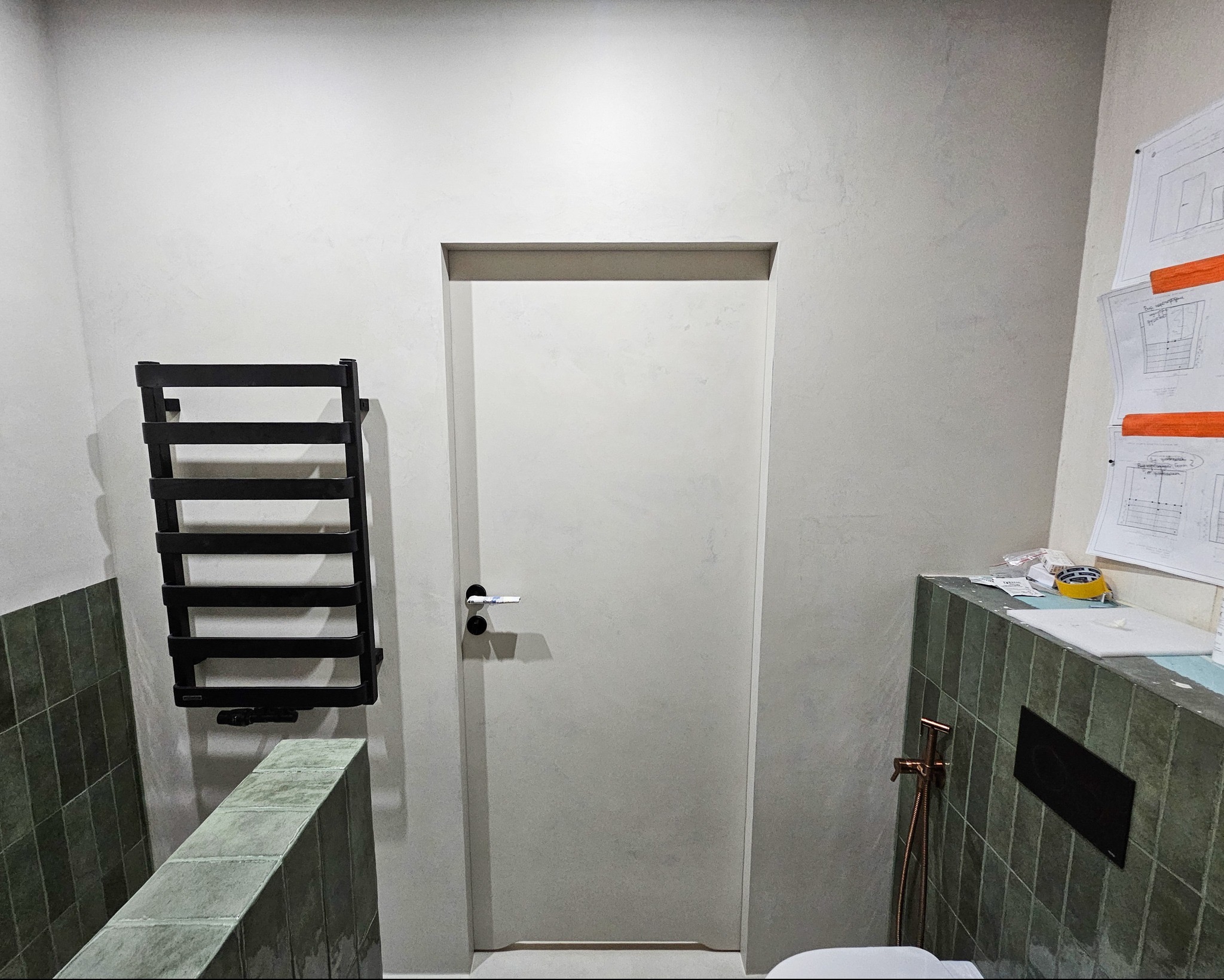 Wnętrze łazienki z minimalistyczną, białą ścianą, drzwiami z czarną klamką, grzejnikiem drabinkowym w kolorze czarnym oraz fragmentem umywalki z zielonymi płytkami. Na ścianie widoczne plany...