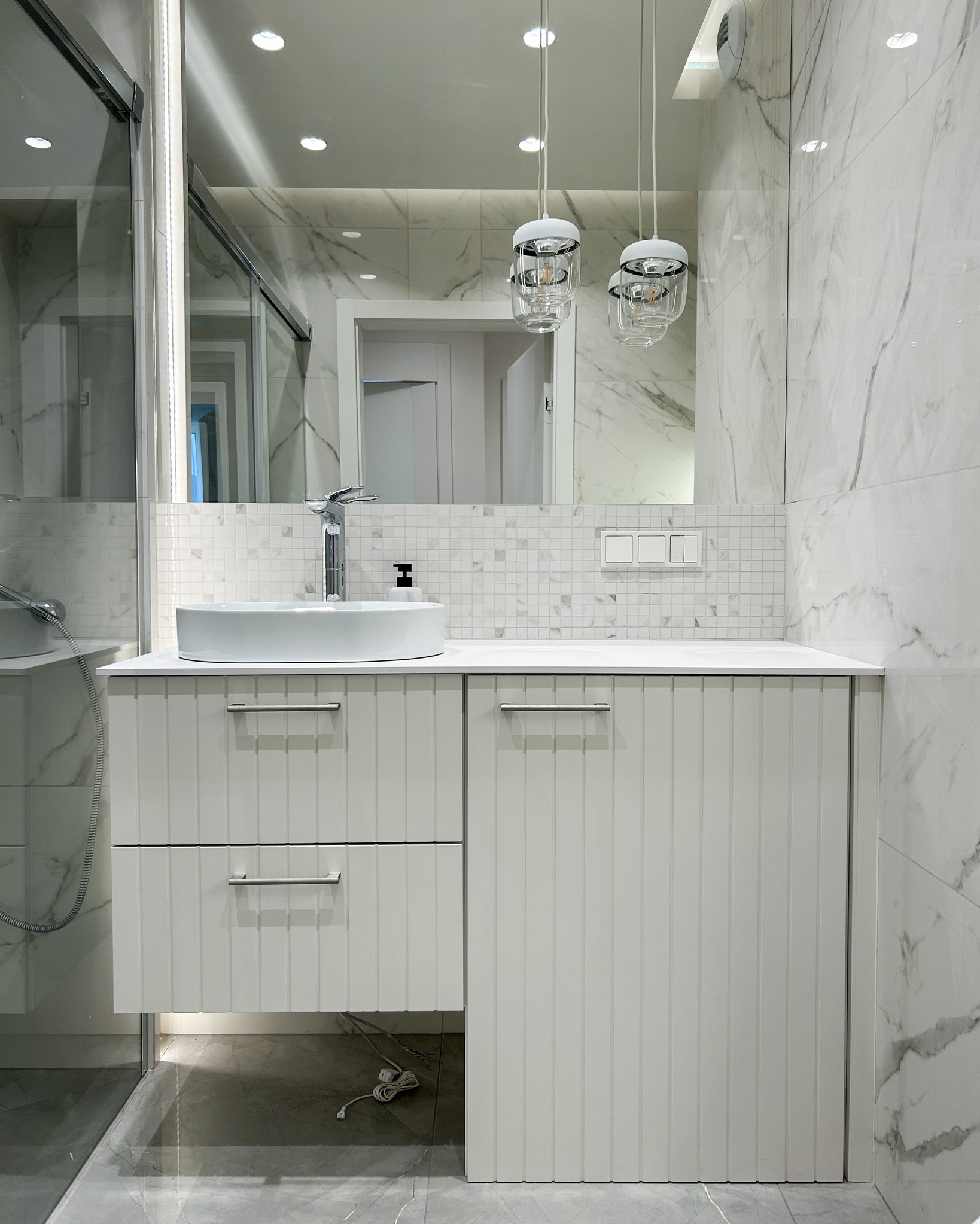 Nowoczesna łazienka z umywalką nablatową, armaturą i szafką z frezowanymi frontami, oświetlona wiszącymi lampami i punktowymi reflektorami, wykończona płytkami imitującymi marmur i mozaiką.
