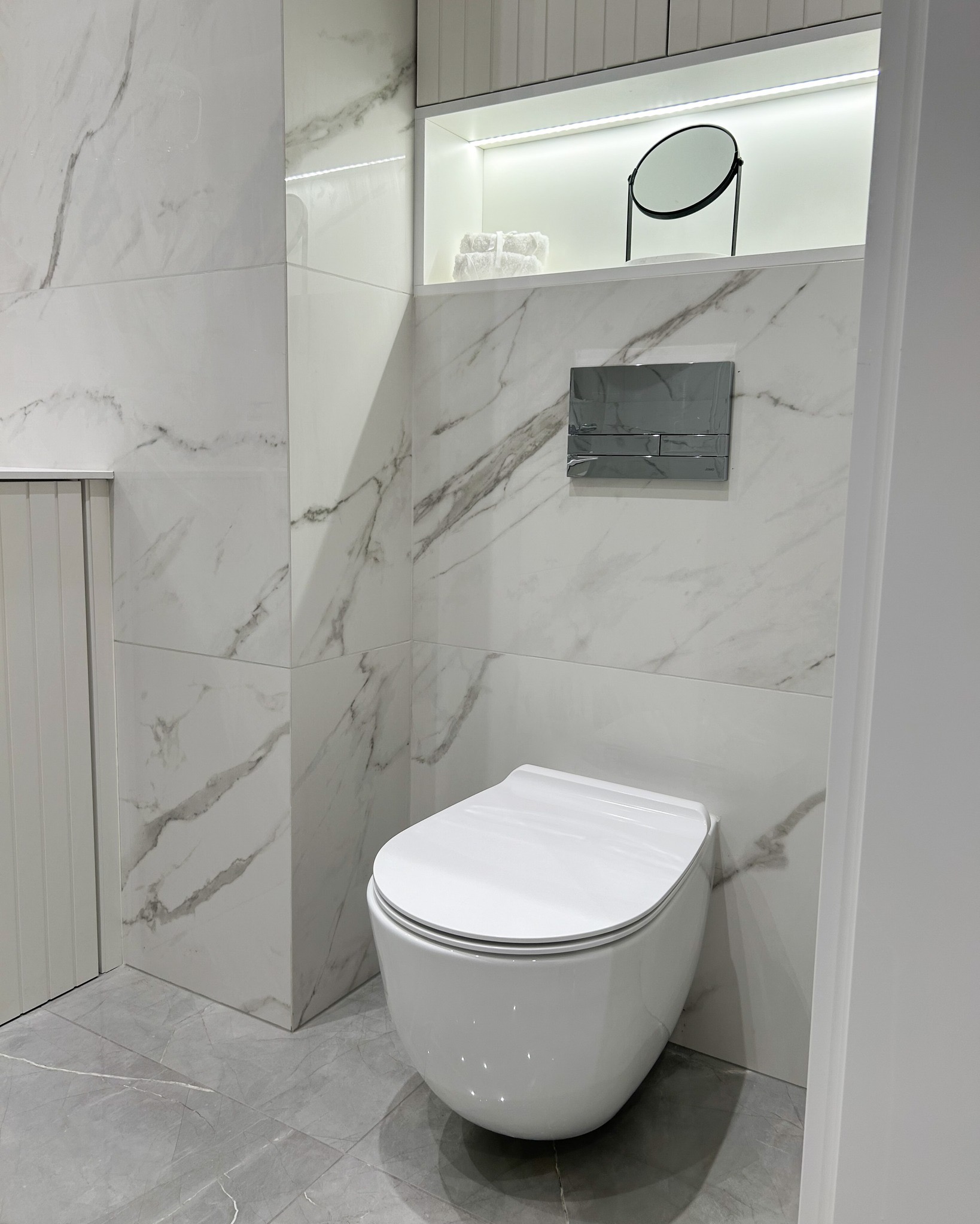 Nowoczesna toaleta podwieszana z chromowanym przyciskiem spłukującym na ścianie wyłożonej białymi płytkami imitującymi marmur. Nad toaletą wnęka z oświetleniem LED, na półce ręczniki i okrągłe...