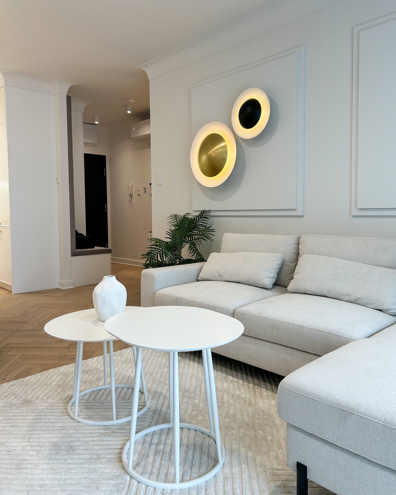 Jasne wnętrze salonu z narożną sofą, dwoma białymi okrągłymi stolikami kawowymi, dekoracyjnymi lampami ściennymi i parkietem ułożonym we wzór jodełki.