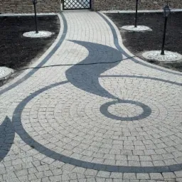 Ogród z ścieżką z kostki brukowej ułożoną w okrągły wzór z ciemnoszarymi akcentami, lampy ogrodowe na trawniku, kamienny murek w tle.