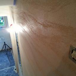 DecorLine - Ściana z efektem dekoracyjnym w odcieniach beżu i brązu, widoczne zabezpieczenia folią malarską, lampa na statywie i niedokończone gniazdko elektryczne.