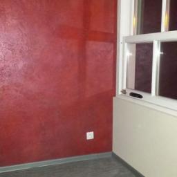 DecorLine - Pokój z czerwoną ścianą o dekoracyjnej fakturze, białe okno z widocznym fragmentem rolety, szara podłoga i gniazdko elektryczne na ścianie.