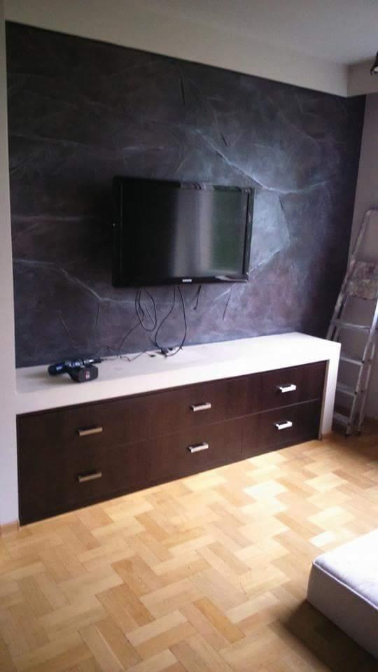 Ściana w salonie wykończona farbą imitującą marmur, podwieszony telewizor, biała komoda z ciemnymi szufladami, parkiet w jodełkę, drabina oparta o ścianę, wiertarka na komodzie.