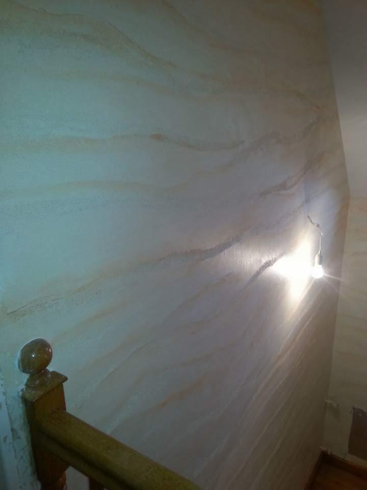 Ściana w korytarzu wykończona dekoracyjną techniką z efektem marmuru w odcieniach beżu i bieli, oświetlona żarówką, widoczna drewniana balustrada schodów.