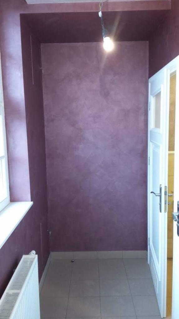 Ściana w małym pokoju wykończona dekoracyjną farbą w odcieniach fioletu i różu, z widoczną fakturą i nierównomiernym rozłożeniem koloru, oświetlona wiszącą żarówką.