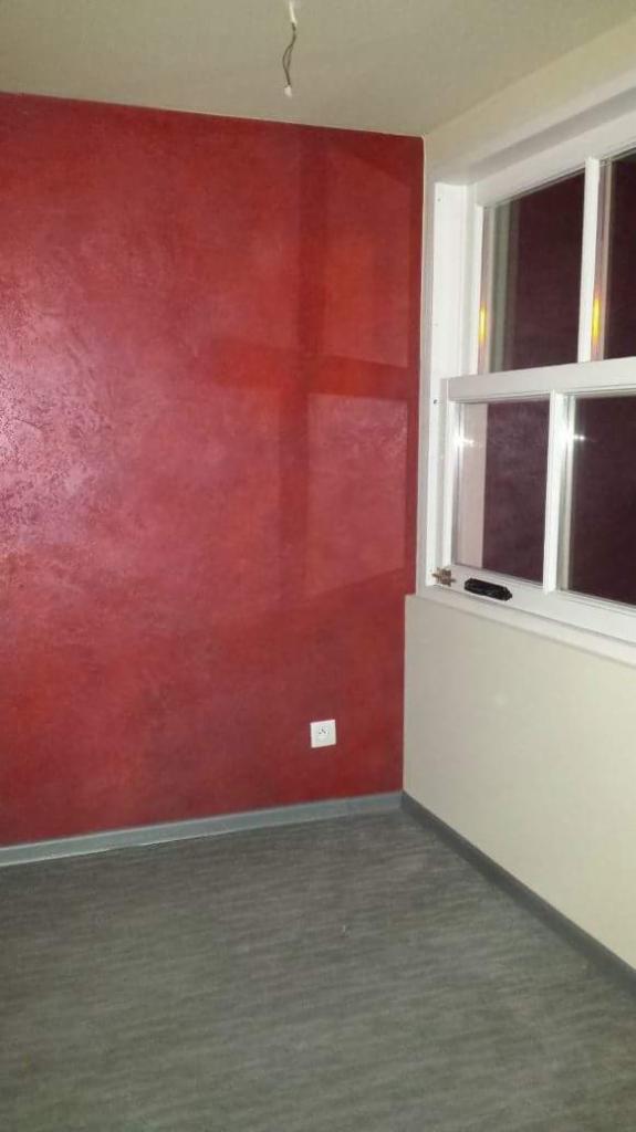 Pokój z czerwoną ścianą o dekoracyjnej fakturze, białe okno z widocznym fragmentem rolety, szara podłoga i gniazdko elektryczne na ścianie.