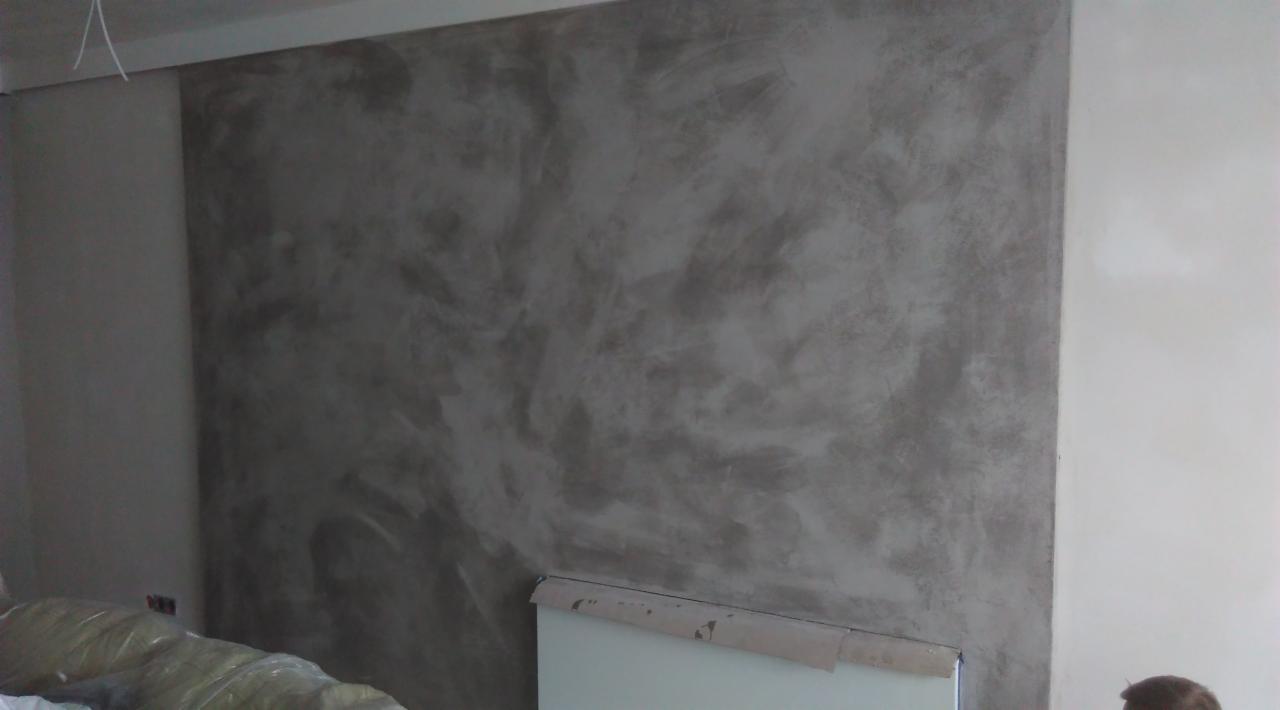 Ściana z dekoracyjną fakturą imitującą beton, sąsiadująca z gładką, pomalowaną na biało ścianą. Widoczne zabezpieczenia podłoża.