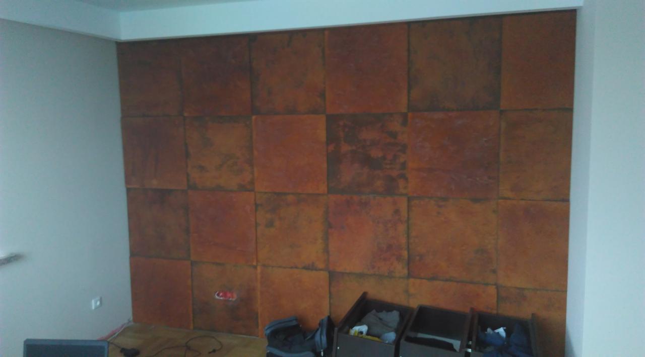 Ściana pokryta panelami imitującymi rdzę, ułożonymi w kwadraty, z widocznym gniazdkiem elektrycznym i fragmentami mebli na dole.