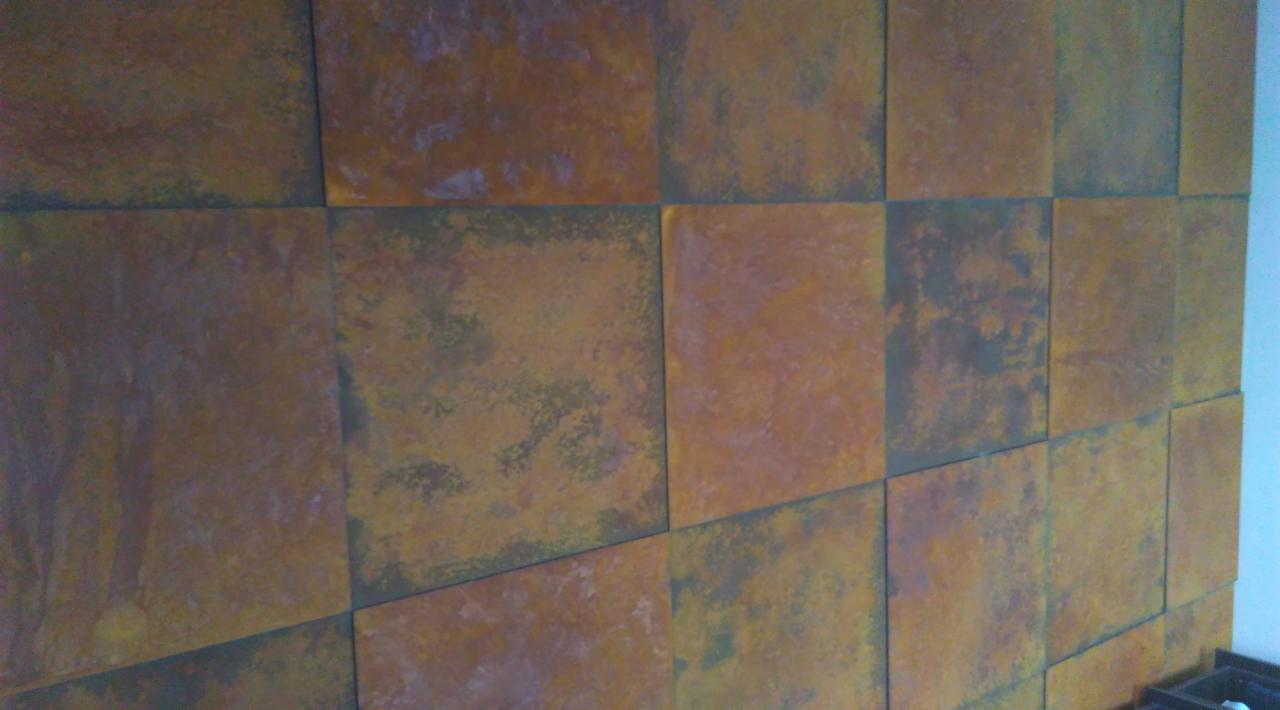 Ściana pokryta kwadratowymi panelami z efektem rdzy, widoczne nieregularne wzory i odcienie brązu, pomarańczy i szarości.
