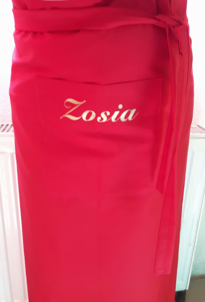 Czerwony fartuch roboczy z kieszenią i złotym napisem 'Zosia', zawiązany w pasie.