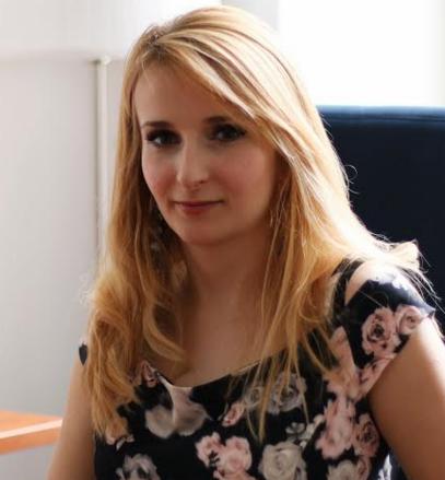 Portret uśmiechniętej kobiety z blond włosami w kwiecistej bluzce, siedzącej przy biurku w biurze.