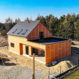 Wznoszony, dwukondygnacyjny budynek z pomarańczową elewacją, widoczne okna dachowe, płaski dach nad garażem i teren przygotowany pod zagospodarowanie.