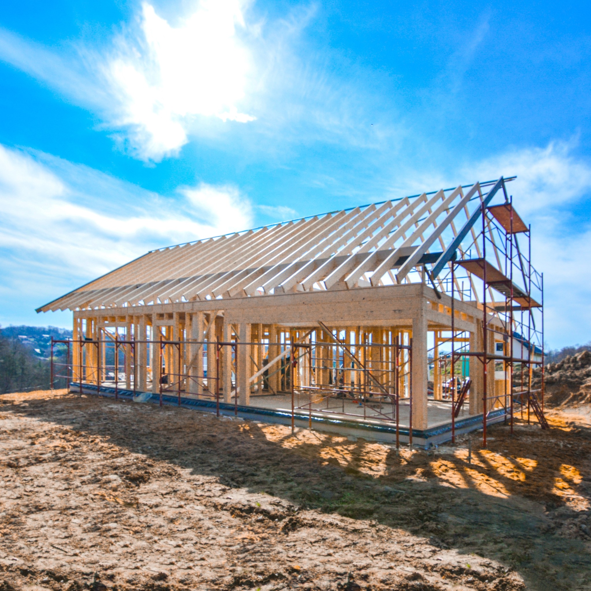 Konstrukcja szkieletowa domu w trakcie budowy, widoczne rusztowania i drewniany szkielet na tle błękitnego nieba z chmurami.