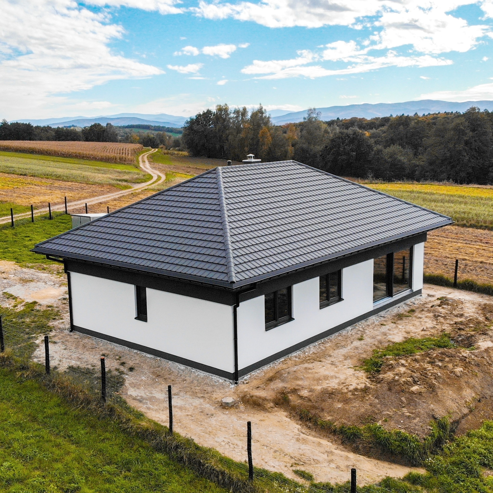 Gotowy, parterowy dom z białymi ścianami i grafitową dachówką, otoczony polami i lasami w oddali, widziany z lotu ptaka.