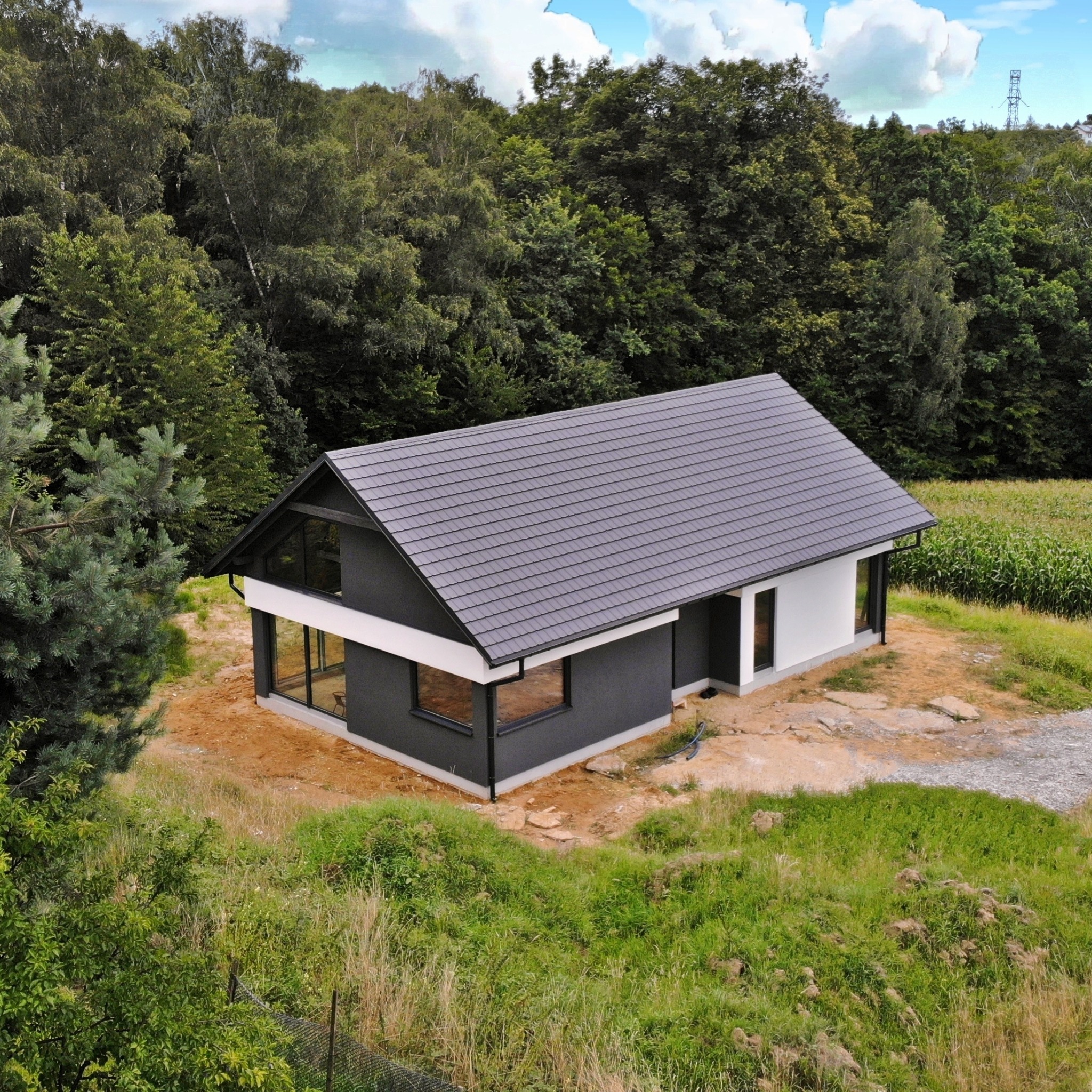Nowoczesny dom z szarą dachówką, czarnymi ramami okiennymi i białymi elementami elewacji, otoczony zielenią i lasem w tle, widziany z góry.