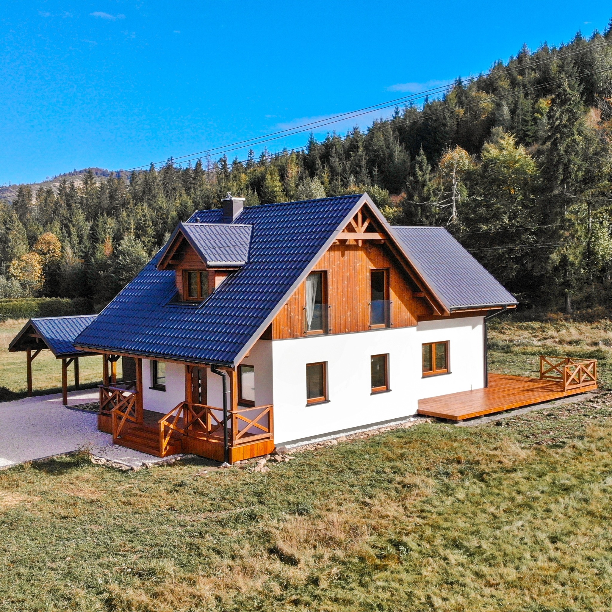 Gotowy dom z białymi ścianami, drewnianą konstrukcją, ciemną dachówką, tarasem i zadaszeniem na tle zalesionego wzgórza.