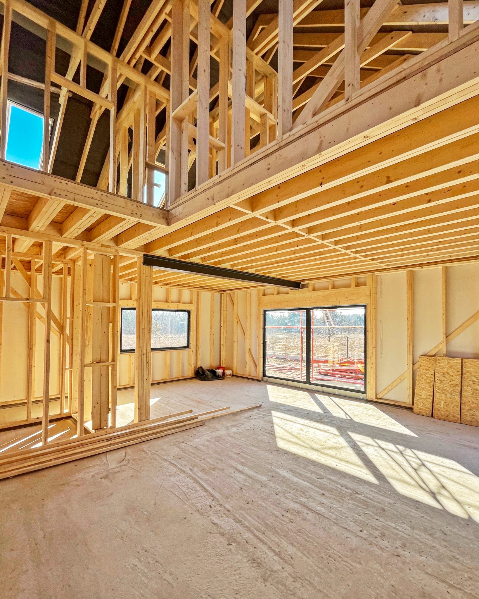 Wnętrze w trakcie budowy, widoczna drewniana konstrukcja szkieletowa, stalowa belka nośna, okna z widokiem na zewnątrz, płyty OSB i surowe podłoże.