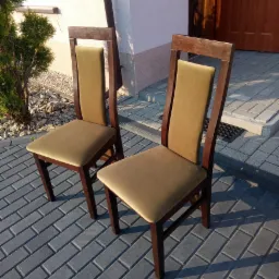 Dwa krzesła z ciemnego drewna z jasnobrązową tapicerką, ustawione na szarym brukowanym chodniku przed domem z zielonym krzewem po lewej stronie i brązowymi drzwiami po prawej.