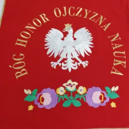Czerwony sztandar z haftowanym białym orłem w koronie, złotymi napisami 'Bóg Honor Ojczyzna Nauka' oraz motywem kwiatowym z frędzlami.