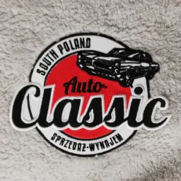 Naszywka z logo 'Auto Classic South Poland' przedstawiająca klasyczny samochód na czerwonym tle, z napisem 'Sprzedaż-Wynajem', umieszczona na miękkim, szarym materiale.