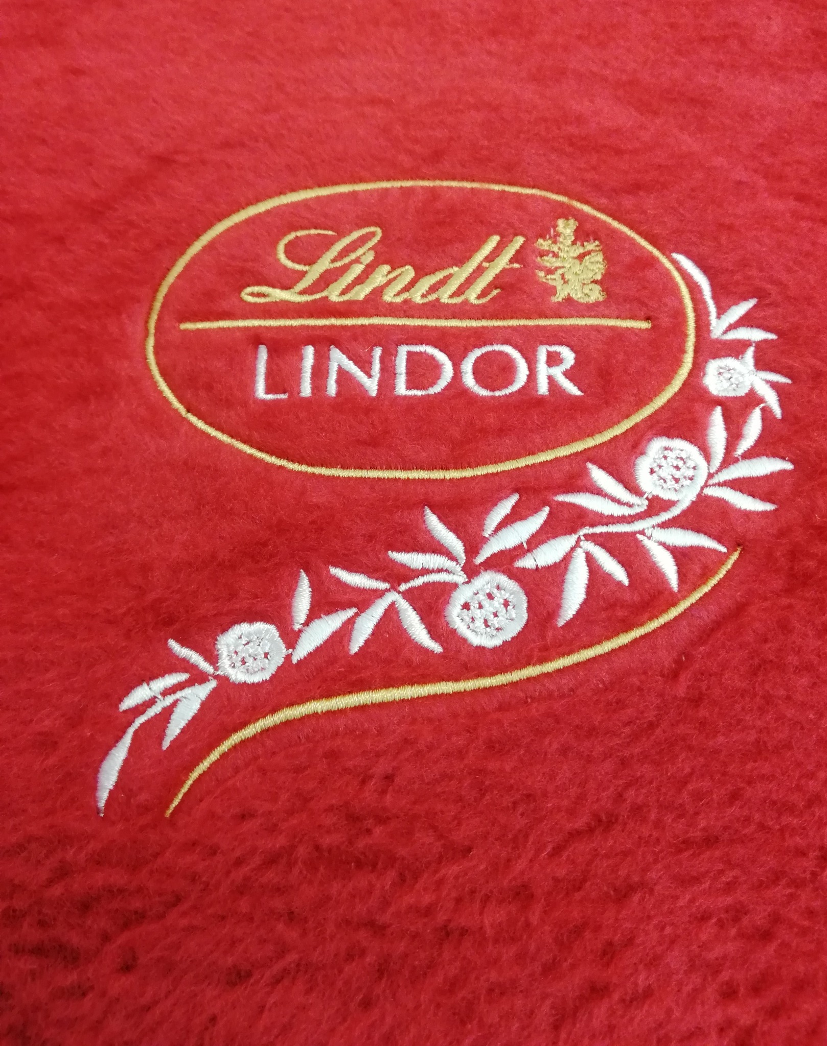 Czerwony, puszysty materiał z wyhaftowanym logo Lindt Lindor w kolorze złotym i białym z motywem roślinnym.