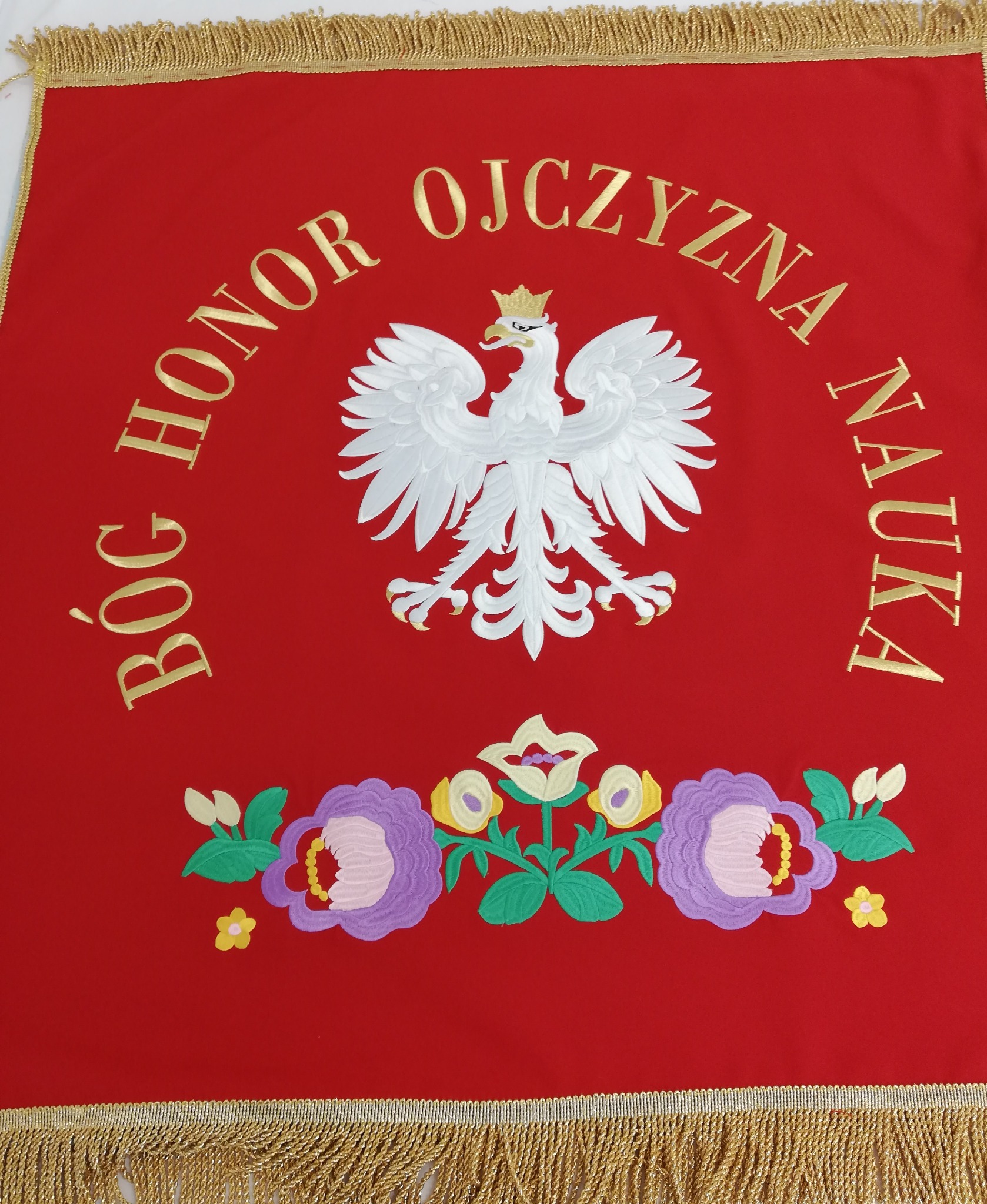 Czerwony sztandar z haftowanym białym orłem w koronie, złotymi napisami 'Bóg Honor Ojczyzna Nauka' oraz motywem kwiatowym z frędzlami.