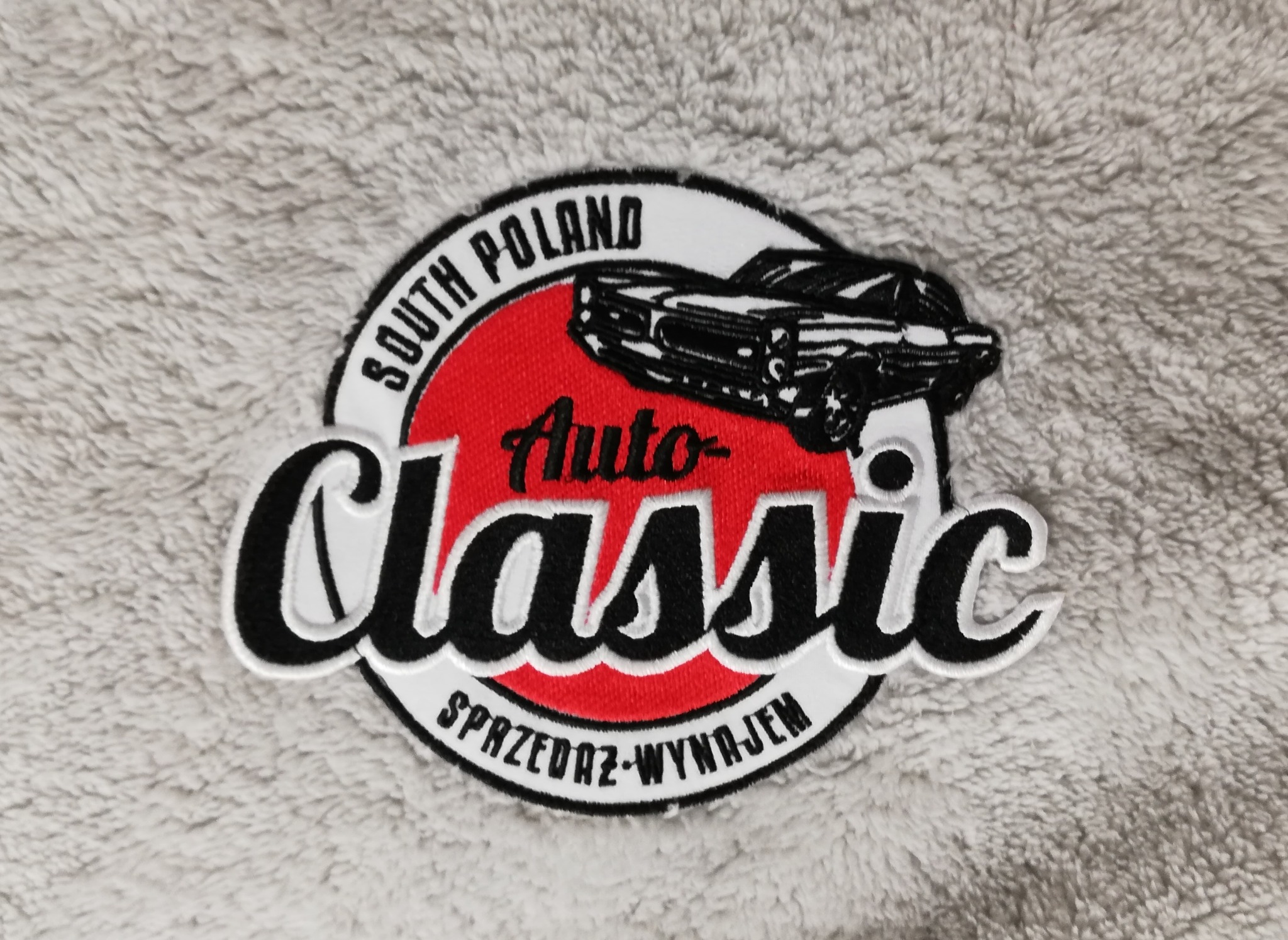 Naszywka z logo 'Auto Classic South Poland' przedstawiająca klasyczny samochód na czerwonym tle, z napisem 'Sprzedaż-Wynajem', umieszczona na miękkim, szarym materiale.