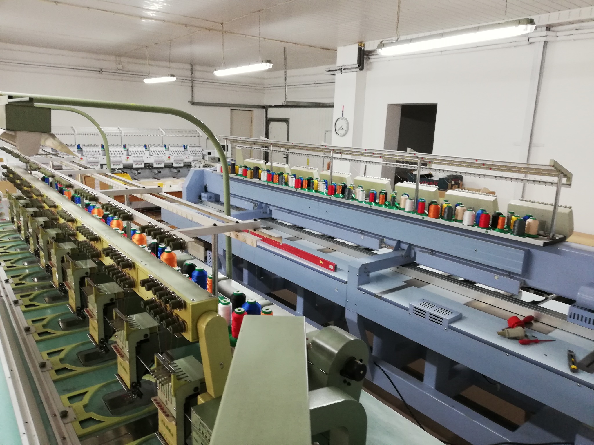 Szeroki kadr hali produkcyjnej z rzędem wielogłowicowych maszyn do haftu przemysłowego, z nawiniętymi kolorowymi nićmi i narzędziami na blacie.